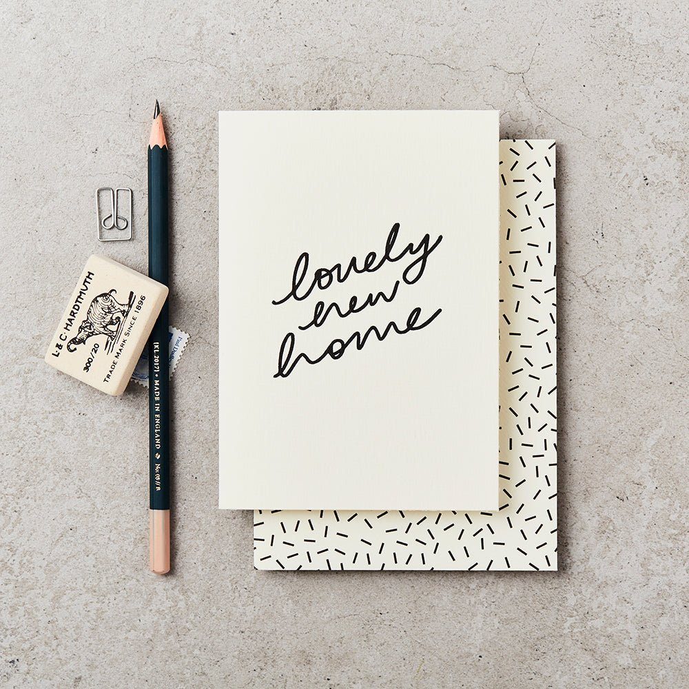 Card with message ' lovely new home' In a handwritten font. Includes black and white envelope with confetti design