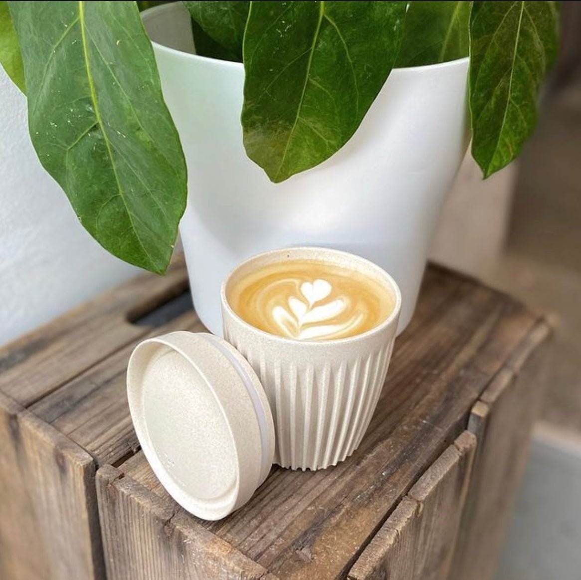 cream coffee cup holding a latte, and stood on a crate, beside a plant pot