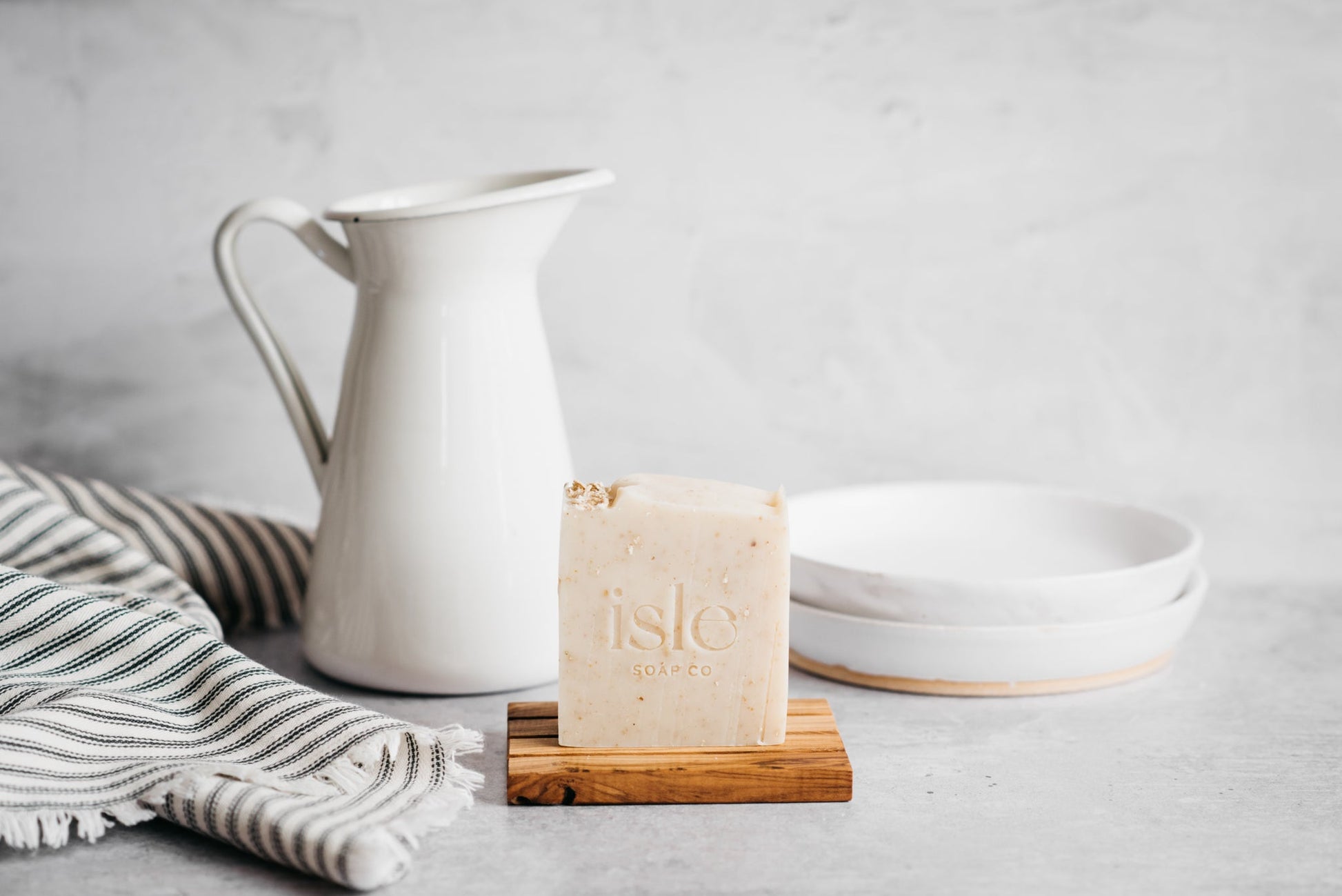 a cream bar of soap standing on a wooden block, with a jug and tea towel behind it.