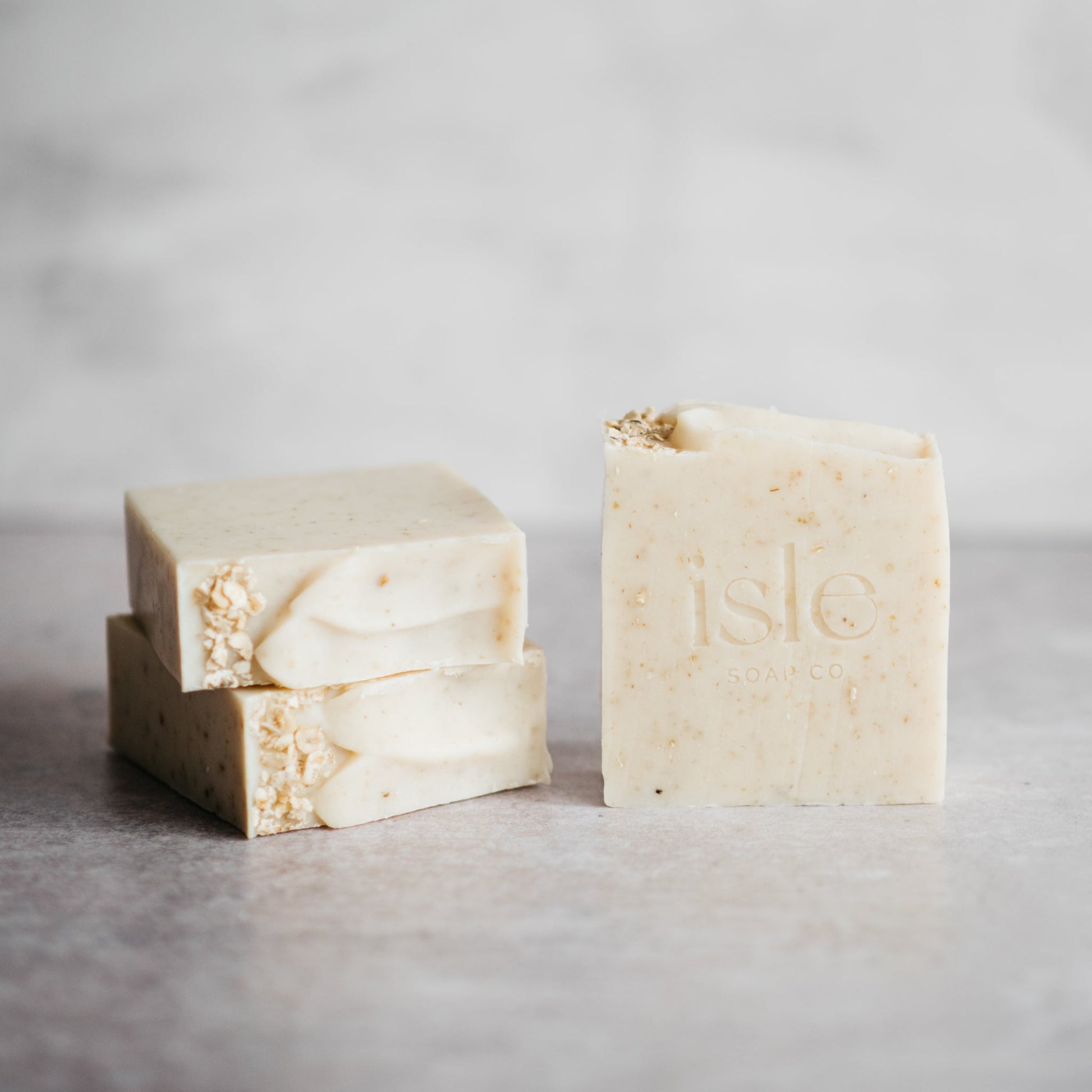 2 square bars of soap stacked on top of each other and a third bar standing up. The soap is cream and shows a scattering of oats on the top. 